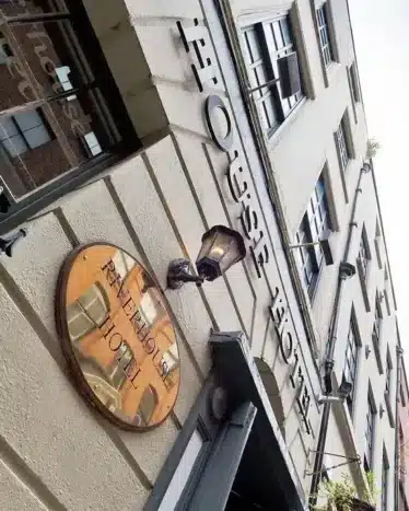 Upward view of a building facade with a round sign reading "Fitzella Hotel" and the words "Fitzhouse Hotel" on the wall above, next to a decorative metal railing.