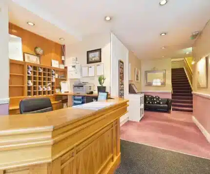 A reception desk with office equipment, cubbyholes, and paperwork in a well-lit hallway. A staircase, sofa, and wall decor are visible in the background.