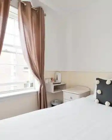 A small, bright bedroom with a double bed, polka dot pillow, brown curtains, a window, a nightstand, and a wall-mounted shelf with a kettle and cups.
