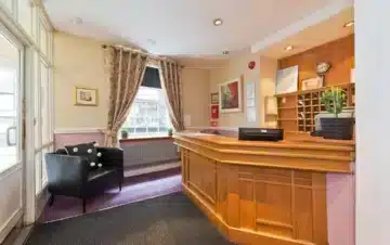 A small hotel reception area with a wooden front desk, a black armchair, a window with curtains, wall art, and a key cubby behind the desk.