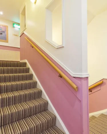 A carpeted staircase with striped pattern, pink and white walls, wooden handrails, emergency exit signs, and a framed picture on the wall.