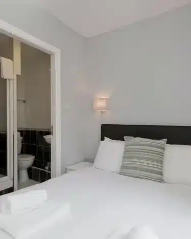 A small bedroom with a double bed, white linens, pillows, and a headboard, adjacent to an en-suite bathroom with a toilet and sink visible through an open door.