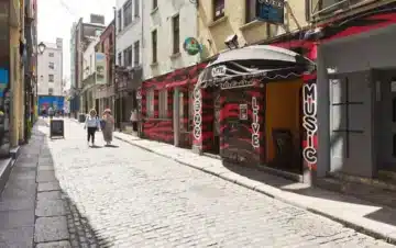 A narrow cobblestone street lined with buildings, including a music venue with a red and black facade; a few people walk in the distance.