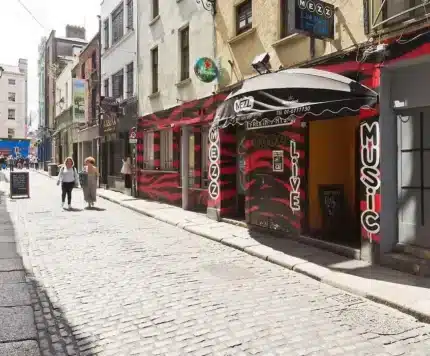 A narrow cobblestone street lined with buildings, including a music venue with a red and black facade; a few people walk in the distance.