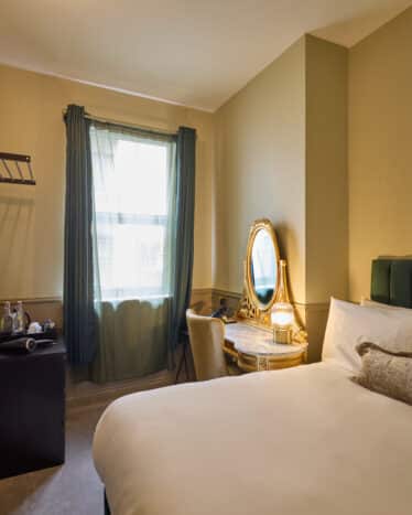 A small hotel room with a double bed, a wall-mounted TV, a desk with a mirror and chair, a window with curtains, and a tea station in the corner.