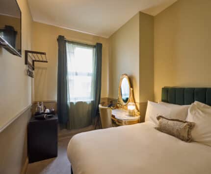 A small hotel room with a double bed, a wall-mounted TV, a desk with a mirror and chair, a window with curtains, and a tea station in the corner.