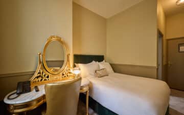 A small hotel room with a double bed, gold vanity table with a mirror, a chair, a telephone, and beige walls.