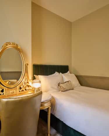 A small hotel room with a double bed, gold vanity table with a mirror, a chair, a telephone, and beige walls.