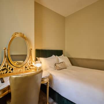 A neatly made bed beside a gold-trimmed vanity with a mirror and chair in a softly lit hotel room with beige walls.