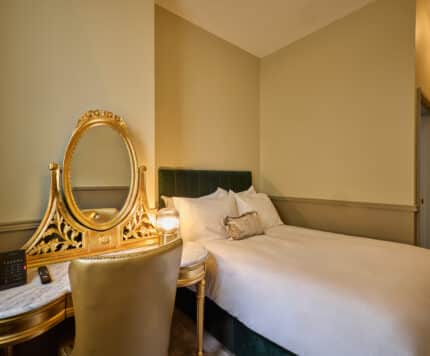 A neatly made bed beside a gold-trimmed vanity with a mirror and chair in a softly lit hotel room with beige walls.