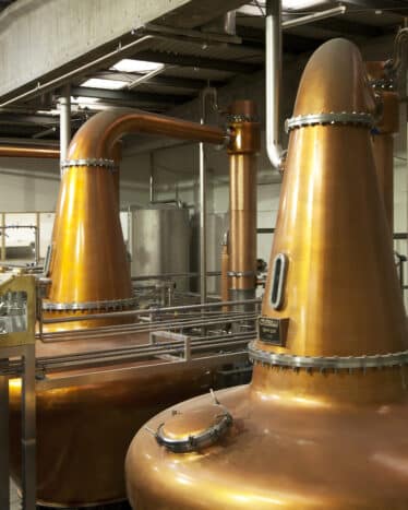 Large copper pot stills inside a whiskey distillery, with pipes and equipment visible; a wall sign reads "Teeling Whiskey" in the background.