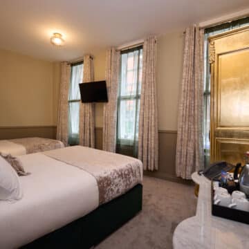Hotel room with a double bed, single bed, gold wardrobe, TV on the wall, and a marble-topped dresser with drinks, cups, and a mirror. Three windows with curtains provide natural light.