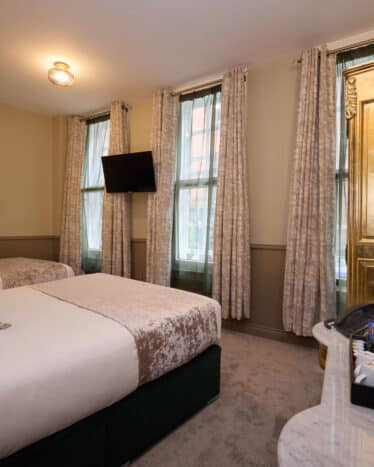 Hotel room with a double bed, single bed, gold wardrobe, TV on the wall, and a marble-topped dresser with drinks, cups, and a mirror. Three windows with curtains provide natural light.