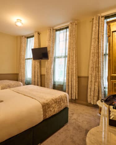 Hotel room with two beds, a wall-mounted TV, tall windows with curtains, and a gold wardrobe with an oval mirror. Bottled drinks and glassware are arranged on a marble table.