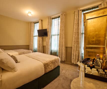 Hotel room with two beds, a wall-mounted TV, tall windows with curtains, and a gold wardrobe with an oval mirror. Bottled drinks and glassware are arranged on a marble table.