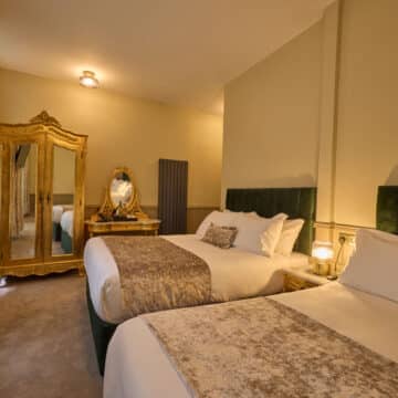 Hotel room with two double beds, ornate gold-framed mirror and wardrobe, green velvet headboards, beige carpet, and patterned curtains.