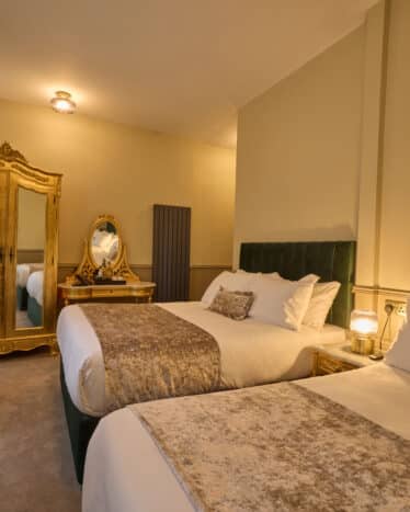 Hotel room with two double beds, ornate gold-framed mirror and wardrobe, green velvet headboards, beige carpet, and patterned curtains.