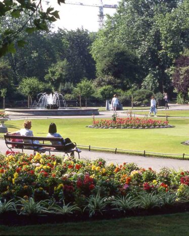 People sit on benches and walk along paths in a landscaped park with colorful flower beds, trimmed lawns, and a central fountain.