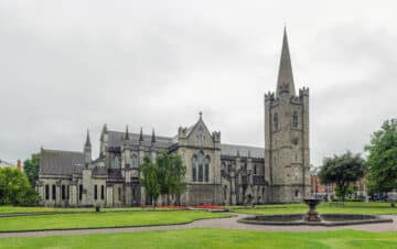 A stone cathedral with a tall spire stands on a grassy lawn with a fountain in front, under an overcast sky.