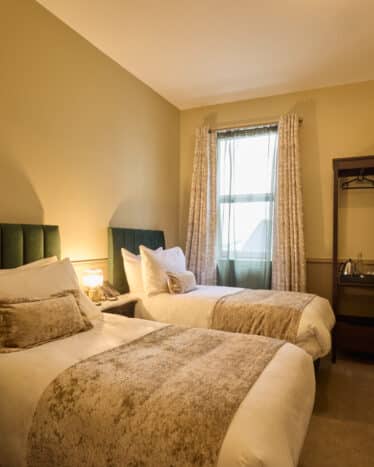 A hotel room features two single beds with plush pillows, a small bedside table with a lamp, a window with curtains, a wall-mounted TV, and a clothes rack.