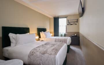 Hotel room with two single beds, white bedding, a wall-mounted TV, a window with curtains, bedside lamps, and a dresser with a kettle and cups.