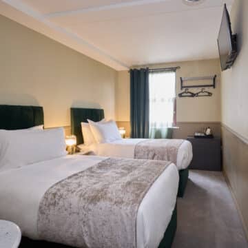 Hotel room with two single beds, white bedding, a wall-mounted TV, a window with curtains, bedside lamps, and a dresser with a kettle and cups.