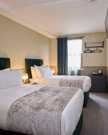 Hotel room with two single beds, white bedding, a wall-mounted TV, a window with curtains, bedside lamps, and a dresser with a kettle and cups.