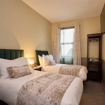A hotel room with two single beds, a nightstand with a lamp, a window with curtains, an open wardrobe, and a wall-mounted TV.