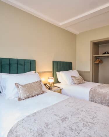 A hotel room with two single beds, green headboards, white bedding, a nightstand with a lamp, an open closet, and a beverage tray on a side table.