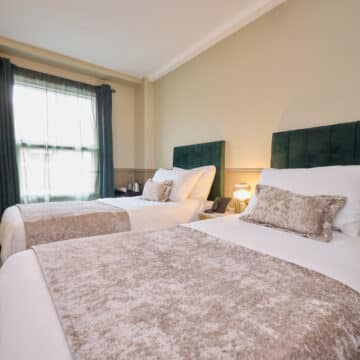 Hotel room with two single beds, green velvet headboards, neutral bedding, a window with dark curtains, wall-mounted TV, and a nightstand with a lamp between the beds.