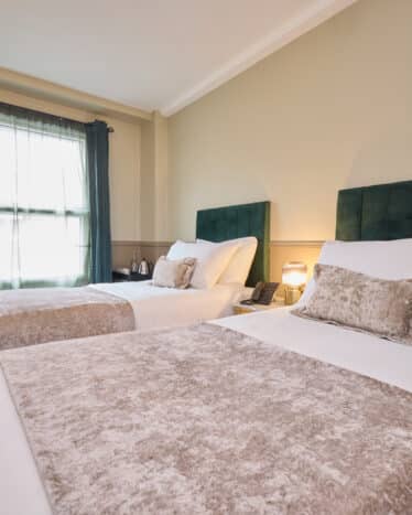 Hotel room with two single beds, green velvet headboards, neutral bedding, a window with dark curtains, wall-mounted TV, and a nightstand with a lamp between the beds.