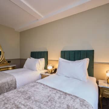 A hotel room with two single beds, green upholstered headboards, gold nightstands with lamps, and a gold vanity with a mirror in the background.
