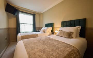 A hotel room with two single beds, each with a green headboard, beige throw, decorative pillow, bedside table, lamp, and a wall-mounted TV near a window with dark curtains.