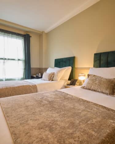 A hotel room with two single beds, each with a green headboard, beige throw, decorative pillow, bedside table, lamp, and a wall-mounted TV near a window with dark curtains.