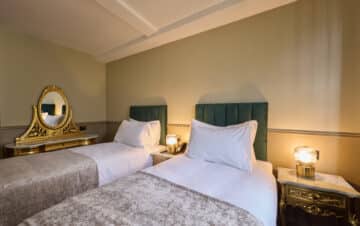 A hotel room with two single beds, green upholstered headboards, a gold vanity table with mirror, and a marble-topped nightstand with a lamp.