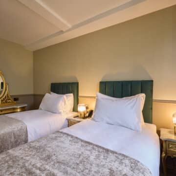 A hotel room with two single beds, green upholstered headboards, a gold vanity table with mirror, and a marble-topped nightstand with a lamp.