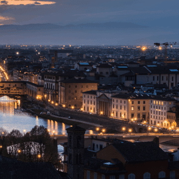 A panoramic view of Florence at dusk, showing the Arno River, illuminated bridges, city buildings, and the Palazzo Vecchio tower against a backdrop of mountains.