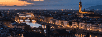 A panoramic view of Florence at dusk, showing the Arno River, illuminated bridges, city buildings, and the Palazzo Vecchio tower against a backdrop of mountains.