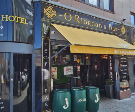 O'Riordan's Bar, a bar and restaurant with yellow signage and awning, stands next to the Arlington Hotel entrance on a city street. Three green Jameson barrels are outside.