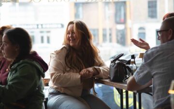 Three people sit at a table in a cafe by a window; one woman in the center is smiling while the others are engaged in conversation.