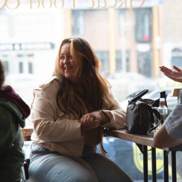 Three people sit at a table in a cafe by a window; one woman in the center is smiling while the others are engaged in conversation.