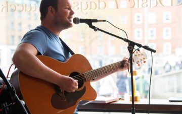 A man in a blue shirt plays an acoustic guitar and sings into a microphone by a large window in a cafe or bar.