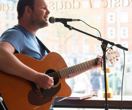 A man in a blue shirt plays an acoustic guitar and sings into a microphone by a large window in a cafe or bar.