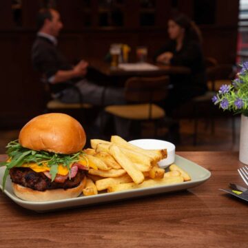 A plate with a burger, fries, and a small cup of sauce on a table in a restaurant, with two people sitting and talking in the background.