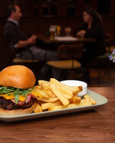 A plate with a burger, fries, and a small cup of sauce on a table in a restaurant, with two people sitting and talking in the background.