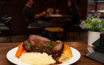 A plate of roast beef with mashed potatoes, carrots, Yorkshire pudding, and gravy on a table; two people are seated and talking in the background.