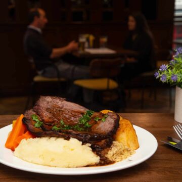 A plate of roast beef with mashed potatoes, carrots, Yorkshire pudding, and gravy on a table; two people are seated and talking in the background.