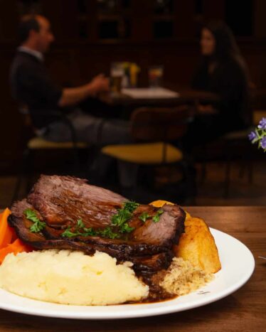 A plate of roast beef with mashed potatoes, carrots, Yorkshire pudding, and gravy on a table; two people are seated and talking in the background.