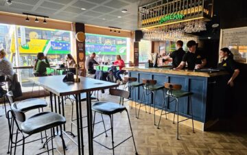 People sit at tables and the bar inside a modern, well-lit pub; staff work behind the bar, and large windows show a bus passing outside.