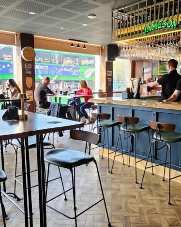 People sit at tables and the bar inside a modern, well-lit pub; staff work behind the bar, and large windows show a bus passing outside.